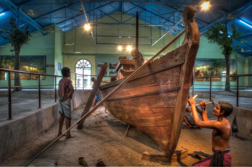 A wooden boat-building display with human figures posed beside a partly built hull.