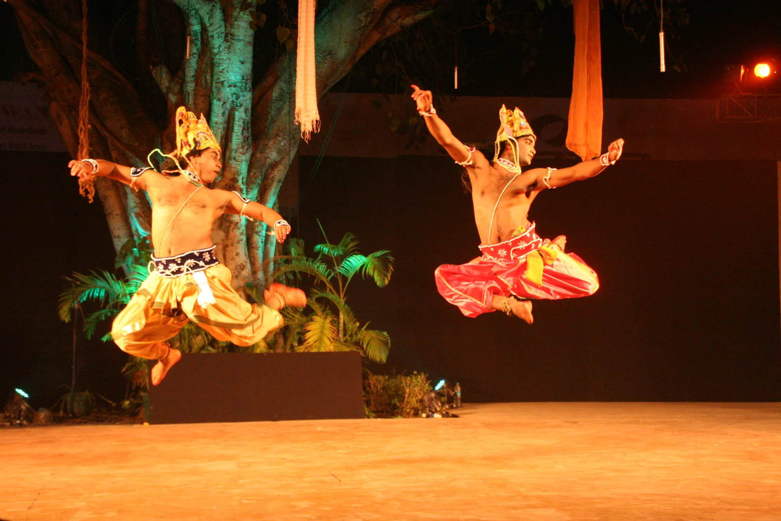 Two unmasked Chhau performers in colourful costumes on a dark stage.