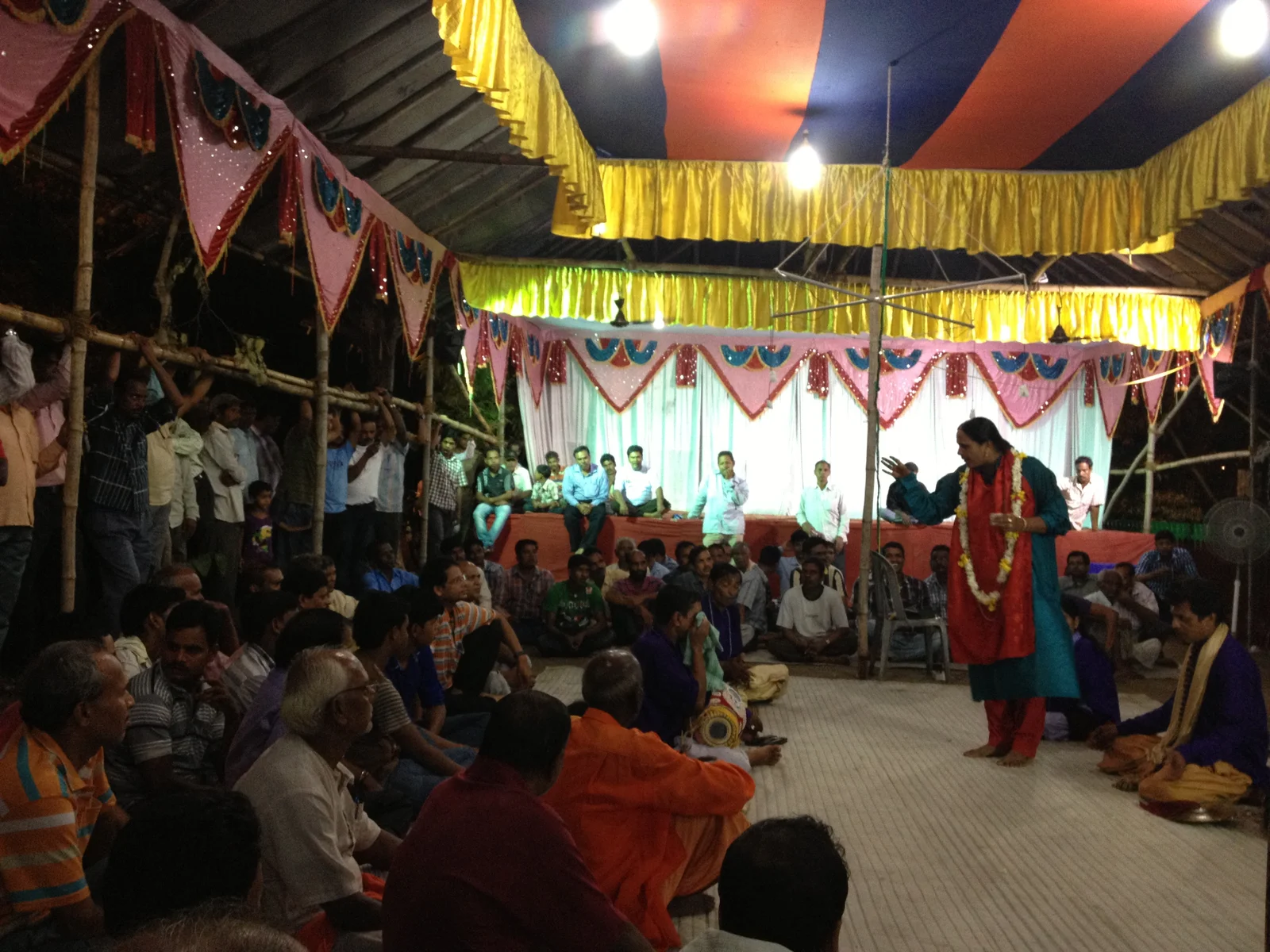 A standing performer at a microphone faces a seated gathering beneath a decorated canopy.